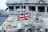 A naval ship with the Union Jack flag prominently displayed, surrounded by various equipment and lifebuoys. The vessel's structure includes windows and metallic surfaces.