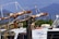 Rusted metal machinery on a docked fishing boat, featuring a winch system and cables. The boat displays a logo with the words 'Wild Caught Seafood.' In the background, there are distant green trees and hazy mountain ranges under a clear blue sky.
