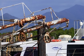 Rusted metal machinery on a docked fishing boat, featuring a winch system and cables. The boat displays a logo with the words 'Wild Caught Seafood.' In the background, there are distant green trees and hazy mountain ranges under a clear blue sky.