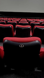 A series of plush red theater seats with black accents are arranged in rows, facing a large blank screen at the front. The seats have numbered plates attached to the backs, with the closest visible seat labeled with the number 6.