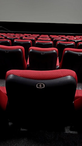 A series of plush red theater seats with black accents are arranged in rows, facing a large blank screen at the front. The seats have numbered plates attached to the backs, with the closest visible seat labeled with the number 6.