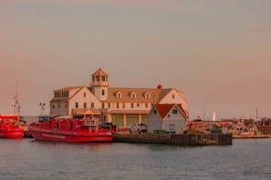 A waterfront scene featuring a large white building with a watchtower and several garage doors, adjacent to smaller structures. Moored along the dock are bright red fire department boats, surrounded by a harbor with various smaller boats and equipment. The sky is overcast with a warm, soft hue casting a serene ambiance over the scene.