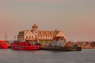 A waterfront scene featuring a large white building with a watchtower and several garage doors, adjacent to smaller structures. Moored along the dock are bright red fire department boats, surrounded by a harbor with various smaller boats and equipment. The sky is overcast with a warm, soft hue casting a serene ambiance over the scene.