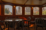 An elegant interior of what appears to be a dining area on a boat or a ship, featuring round tables with chairs and large windows offering a view of a cityscape with skyscrapers and greenery.