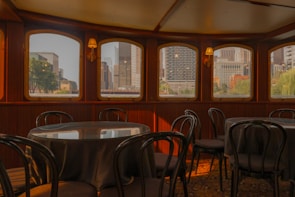 An elegant interior of what appears to be a dining area on a boat or a ship, featuring round tables with chairs and large windows offering a view of a cityscape with skyscrapers and greenery.