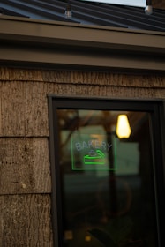 A rustic wooden exterior features a window displaying a glowing green neon sign with the word 'Bakery' and an image of a pastry or cake. A warm light is visible inside the building, creating a cozy, inviting atmosphere.