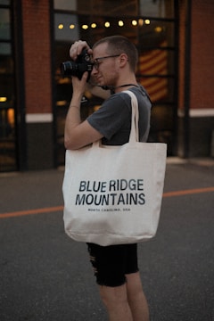 A person with short hair is using a camera to take a photograph. They are wearing glasses, a dark t-shirt, and carrying a white tote bag with the text 'Blue Ridge Mountains, North Carolina, USA'. The background features a building with large windows and warm lighting.