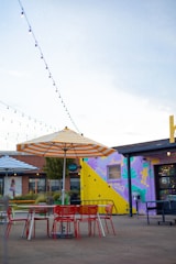 A vibrant outdoor patio area featuring red tables and chairs under large umbrellas with colorful stripes. A mural with abstract designs in shades of purple, yellow, and blue decorates a wall of a nearby building. String lights are hanging above, creating a festive and lively atmosphere near a building with large windows.
