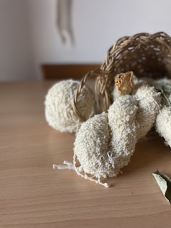 A wooden table holds a woven basket turned on its side, spilling out a few items. There are textured, cream-colored balls and a skein of yarn. Dry leaves are also present nearby, contributing to a natural and rustic arrangement.