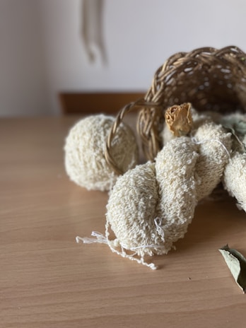 A wooden table holds a woven basket turned on its side, spilling out a few items. There are textured, cream-colored balls and a skein of yarn. Dry leaves are also present nearby, contributing to a natural and rustic arrangement.