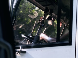 View through the window of a vehicle cabin focused on the steering wheel, dashboard, and an arm operating controls. The glass reflects natural surroundings and features interior elements like mounted fans and equipment.