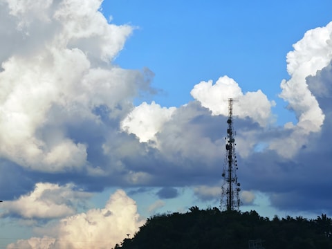 A tall communication tower is silhouetted against a background of large, fluffy clouds in varying shades of white and gray. The sky is a vibrant blue, creating a striking contrast with the darker clouds. The tower is situated on a hill covered with trees, which can be seen at the bottom of the image.