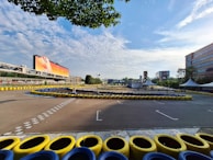 A go-kart track surrounded by yellow and blue tire barriers is visible under a partly cloudy sky. The track is empty and situated near a large advertisement billboard. Buildings and trees surround the area, with a checkered tower and tents visible in the distance.