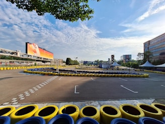 A go-kart track surrounded by yellow and blue tire barriers is visible under a partly cloudy sky. The track is empty and situated near a large advertisement billboard. Buildings and trees surround the area, with a checkered tower and tents visible in the distance.
