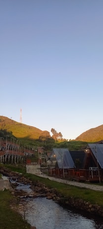 A serene landscape featuring a small stream running through a lush green area. There are modern triangle-roofed structures along the side of the stream, surrounded by well-maintained grass and hills in the background. A footbridge crosses the stream and a few trees are sprinkled across the scene. The sky is clear and blue, with a hilltop visible in the distance.