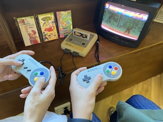 Two people are playing a retro video game on a Super Nintendo console. They are holding SNES controllers, and there is a vintage CRT television displaying a fighting game. Several game cartridges with colorful covers are displayed in the background.