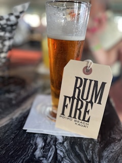 A tall, frothy glass of beer rests on a dark, marbled surface with a paper coaster beneath it. Attached to the glass is a tag with bold black letters reading 'Rum Fire Poipu Beach Kauai'. The background appears to be blurry, suggesting a lively or busy atmosphere, possibly a bar or restaurant.
