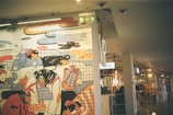 A colorful mural portrays various whimsical characters engaged in playful activities, with abstract and doodle-like artistic elements. In the background, there is an indoor shopping environment with signs and a visible retail layout.