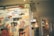 A colorful mural portrays various whimsical characters engaged in playful activities, with abstract and doodle-like artistic elements. In the background, there is an indoor shopping environment with signs and a visible retail layout.