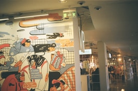 A colorful mural portrays various whimsical characters engaged in playful activities, with abstract and doodle-like artistic elements. In the background, there is an indoor shopping environment with signs and a visible retail layout.