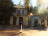 A historic stone building with the sign 'Placerville Soda Works' is next to a colorful shop with a sign that reads 'Empress'. The building has large windows and is surrounded by tall trees. Sunlight filters through the trees, creating lens flares and a warm glow in the image.