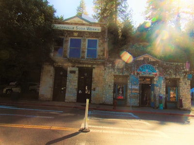 A historic stone building with the sign 'Placerville Soda Works' is next to a colorful shop with a sign that reads 'Empress'. The building has large windows and is surrounded by tall trees. Sunlight filters through the trees, creating lens flares and a warm glow in the image.