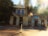 A historic stone building with the sign 'Placerville Soda Works' is next to a colorful shop with a sign that reads 'Empress'. The building has large windows and is surrounded by tall trees. Sunlight filters through the trees, creating lens flares and a warm glow in the image.