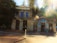 A historic stone building with the sign 'Placerville Soda Works' is next to a colorful shop with a sign that reads 'Empress'. The building has large windows and is surrounded by tall trees. Sunlight filters through the trees, creating lens flares and a warm glow in the image.