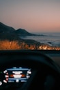 A scenic coastal view at sunset or sunrise is visible, with mountains in the background and the ocean stretching out to the right. The foreground shows the dashboard of a car, hinting at a road trip or scenic drive. The warm glow of headlights illuminates dry grass near the edge of the road.