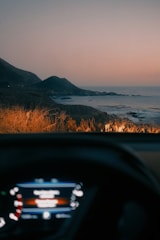 A scenic coastal view at sunset or sunrise is visible, with mountains in the background and the ocean stretching out to the right. The foreground shows the dashboard of a car, hinting at a road trip or scenic drive. The warm glow of headlights illuminates dry grass near the edge of the road.