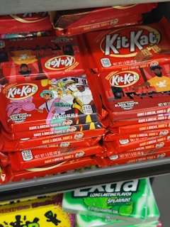 Stacks of KitKat chocolate bars with colorful packaging sit on a shelf. The design on the wrappers includes illustrations of musicians and vibrant scenes, possibly related to New Orleans. Below them, packs of Extra spearmint gum are visible.