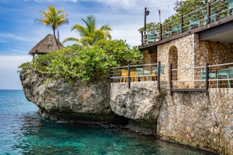 A scenic coastal view featuring a rustic stone building with a balcony overlooking the ocean. The building is adorned with several green and yellow chairs, and a lush green shrubbery atop a large rock formation can be seen. Tall palm trees rise above the structure, adding a tropical touch to the setting. The clear, blue water gently laps at the base of the rocky outcrop.