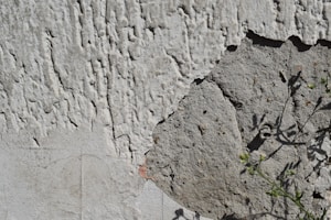 A close-up view of a weathered wall with peeling stucco or plaster. The texture is rough and uneven, with visible cracks and exposed concrete underneath. Shadows of small plants can be seen on the surface, indicating nearby vegetation.