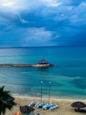 A serene beachfront scene with calm turquoise waters under a cloudy sky. Three catamarans are parked on the sandy shore, and a small wooden gazebo sits on a rocky outcrop extending into the sea. Palm trees and thatched umbrellas add to the tropical atmosphere.