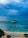 A serene beachfront scene with calm turquoise waters under a cloudy sky. Three catamarans are parked on the sandy shore, and a small wooden gazebo sits on a rocky outcrop extending into the sea. Palm trees and thatched umbrellas add to the tropical atmosphere.