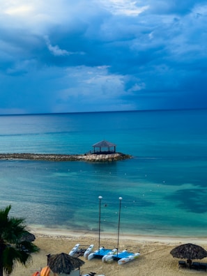 A serene beachfront scene with calm turquoise waters under a cloudy sky. Three catamarans are parked on the sandy shore, and a small wooden gazebo sits on a rocky outcrop extending into the sea. Palm trees and thatched umbrellas add to the tropical atmosphere.