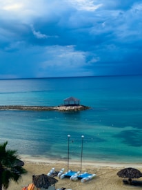 A serene beachfront scene with calm turquoise waters under a cloudy sky. Three catamarans are parked on the sandy shore, and a small wooden gazebo sits on a rocky outcrop extending into the sea. Palm trees and thatched umbrellas add to the tropical atmosphere.