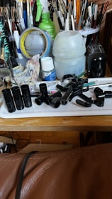 A cluttered workspace with a variety of art supplies, including paintbrushes of different sizes and colors, a roll of masking tape, containers, a spray bottle, a rag with paint stains, and several black plastic items on a tray. The setting conveys a busy and well-used area for creating art.