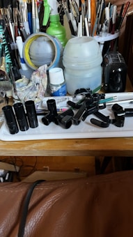 A cluttered workspace with a variety of art supplies, including paintbrushes of different sizes and colors, a roll of masking tape, containers, a spray bottle, a rag with paint stains, and several black plastic items on a tray. The setting conveys a busy and well-used area for creating art.