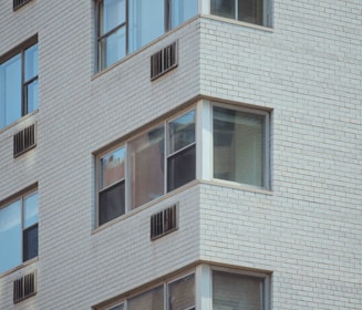 A close-up view of a multi-story residential building facade with white brick walls and several windows. Some windows have air conditioning units installed below them.