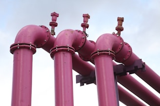 Four large pink industrial pipes extend upward, each with a valve on top, set against a light blue sky.