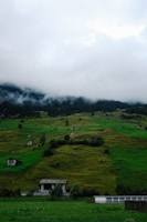 A lush, green hill dotted with several small houses and cabins. The sky above is cloudy and overcast, with patches of mist lingering around the top of the hill. Tall trees and bushes are scattered across the landscape, adding an element of natural beauty to the scene.