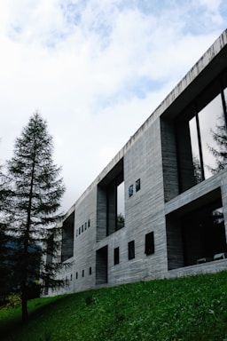 A modern cement building with large rectangular windows, set on a grassy slope. The façade features a minimalist design with simple lines and a textured finish. Tall trees are positioned on the left, and a cloudy, yet bright, sky fills the background.