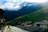 A picturesque village nestled beside a gently flowing river with quaint houses, green rolling hills, and towering mountains in the background. The mountains are partially covered by misty clouds, and sunlight softly illuminates the scene, creating a serene and idyllic atmosphere.
