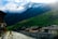 A picturesque village nestled beside a gently flowing river with quaint houses, green rolling hills, and towering mountains in the background. The mountains are partially covered by misty clouds, and sunlight softly illuminates the scene, creating a serene and idyllic atmosphere.