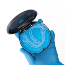 A gloved hand holds a black case with a blue interior, containing a clear dental aligner. The aligner is transparent and designed to fit over teeth, indicating orthodontic use.