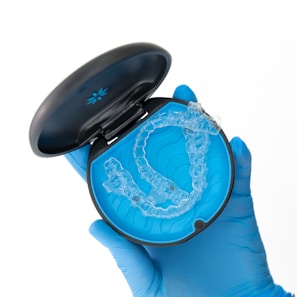 A gloved hand holds a black case with a blue interior, containing a clear dental aligner. The aligner is transparent and designed to fit over teeth, indicating orthodontic use.