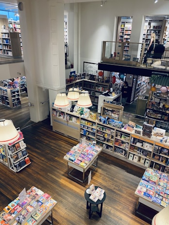 A spacious and brightly lit bookstore features wooden flooring and multiple shelves filled with books and games. The lower level showcases tables with colorful books neatly arranged, while the upper level includes additional shelves and people browsing. Illumination is provided by large ceiling lamps, contributing to a cozy and inviting atmosphere.