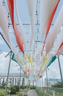 Colorful fabric strips hang from a metal frame with built-in light bulbs, creating a vibrant canopy over a walkway. The scene is set against a blue sky with fluffy clouds and surrounded by greenery and solar panels.