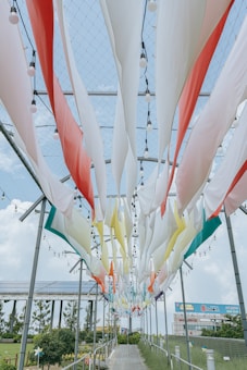 Colorful fabric strips hang from a metal frame with built-in light bulbs, creating a vibrant canopy over a walkway. The scene is set against a blue sky with fluffy clouds and surrounded by greenery and solar panels.